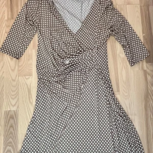 Brun prickig omlottklänning - Snygg brun omlottklänning med vita prickar och trekvartsärmar. Perfekt för en stilren look med en v-ringad design som ger en elegant touch.