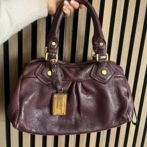 Marc Jacobs väska  - Marc Jacobs classic q baby groove med guldiga detaljer. Väskan är lila/vinröd. Axelremband ingår. Den är ca 30 cm bred och är i den lilla modellen. Bra skick. Finns lite slitningar, se bilder. Första bilden är utan blixt resten är med blixt. Fler bilder finns och svarar gärna på frågor 💜