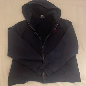 Mörkblå hoodie från Ralph Lauren -  Xl barn som motsvarar S-M. Säljer en snygg mörkblå hoodie från Ralph Lauren med dragkedja och det klassiska röda broderade logotypen på bröstet. Perfekt för en avslappnad stil. Hoodien har långa ärmar och en bekväm passform. 