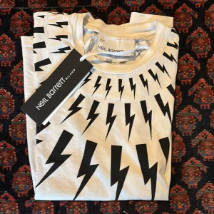 Vit t-shirt med blixtmönster från Neil Barrett - Snygg vit t-shirt från Neil Barrett med ett coolt svart blixtmönster över bröstet. Perfekt för att ge din outfit en edgy touch. Kortärmad och i mjukt material för maximal komfort.