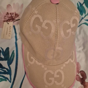 Beige och rosa keps från Gucci - Snygg beige keps från Gucci med rosa detaljer och det ikoniska GG-mönstret. Kepsen har en justerbar rem i rosa läder baktill för perfekt passform. Perfekt för att ge din outfit en lyxig touch.