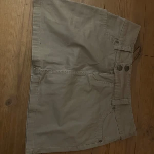 Beige kort kjol med fickor - Snygg beige kortkjol med praktiska fickor fram och bak. Kjolen har en klassisk design med två knappar i midjan och bälteshällor för extra stil. Perfekt för en avslappnad look.