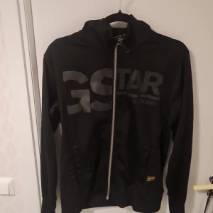 Svart jacka från G-Star RAW - Säljer en svart jacka från G-Star RAW med dragkedja och Jackan har ett stort tryck på framsidan och en liten etikett med logotypen. Perfekt för en casual look.