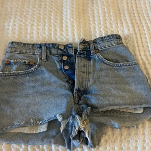 Blå jeansshorts från Zara - Säljer ett par snygga blå jeansshorts från Zara. De har en klassisk femficksdesign med knappar framtill och fransiga kanter för en avslappnad look. Perfekta för sommardagar!