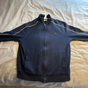 Mörkblå tröja från Lacoste - Säljer en stilren mörkblå tröja från Lacoste med dragkedja och vita detaljer längs ärmarna. Jackan har en hög krage och två fickor framtill. Perfekt för en sportig look eller när man är ute med vänner o familj
