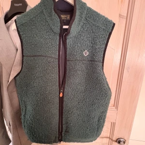 Grön Morris fleeceväst med dragkedja mo - Säljer en mysig grön fleeceväst med dragkedja och en liten broderad detalj på bröstet. Perfekt för kyliga dagar och lätt att matcha med andra plagg. Västen har en normal passform och är idealisk för både höst och vår.