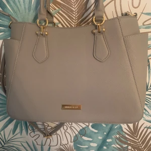 Beige axelväska med gulddetaljer - Säljer en  beige axelväska med eleganta gulddetaljer. Väskan har en praktisk dragkedja och är perfekt för att bära dina dagliga nödvändigheter. Den har många fickor ås man rymmer mycket💗Obs! Om ni ser likadana plagg som jag säljer är det förmodligen mitt gamla konto som inte längre fungerar!