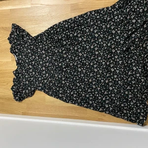 Svart blommig klänning - Charmig svart klänning med ett sött blommönster i vitt och grönt. Klänningen har korta ärmar och en volangdetalj vid fållen 🌸🌺🌼 Varan är rätt använd men ändå i väldigt bra skick. Skulle säga att storleken är omkring xs/s 💗💗