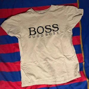 Vit t-shirt från Hugo Boss - Säljer en stilren vit t-shirt från Hugo Boss med stor svart logga på framsidan. Perfekt för en avslappnad look. T-shirten har en klassisk rund hals och korta ärmar. Oanvänd. Säljer pga inte använder den.