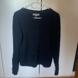 Svart kofta från Basiqwear - Snygg svart kofta från Basiqwear med knappar framtill. Den har ett diskret ribbat mönster och långa ärmar. Perfekt för lager-på-lager-stil eller som ett enkelt basplagg i garderoben.