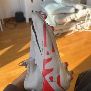 Vita och röda fotbollsskor från Nike - Snygga fotbollsskor från Nike i vitt med röda och svarta detaljer. Skorna har en strömlinjeformad design med dobbar för optimalt grepp på planen. Perfekta för fotbollsspelare som vill ha stil och funktionalitet.