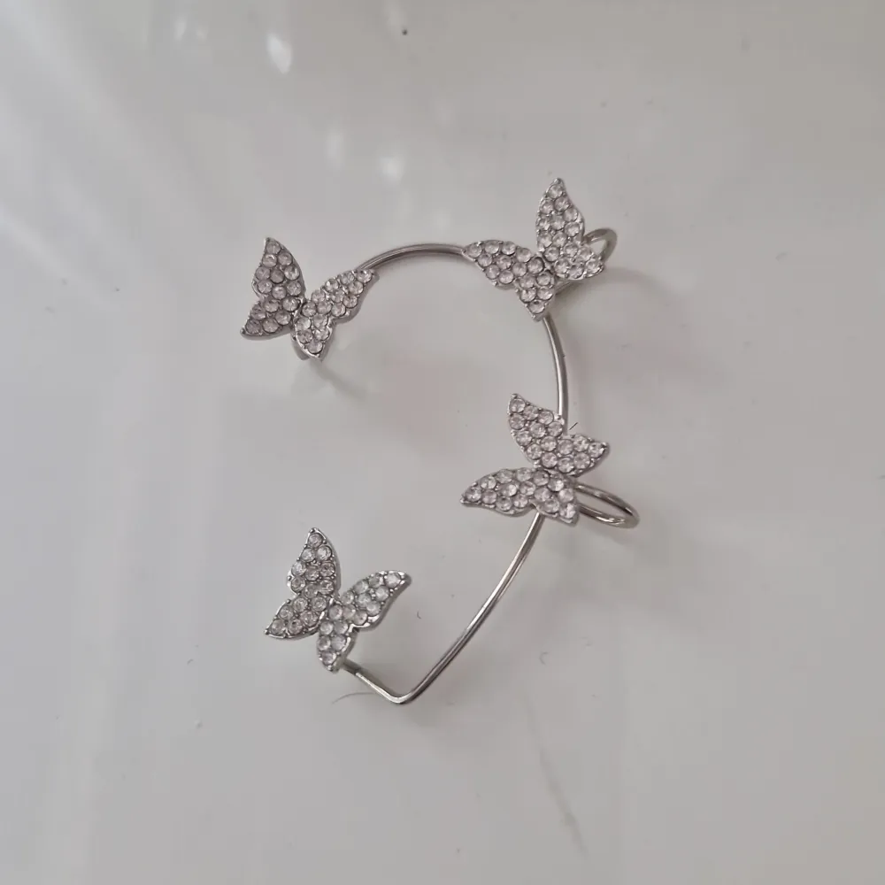 Elegant silverfärgad accessoar dekorerat med glittrande fjärilar. Perfekt för att lägga till en touch av glamour till din outfit. Den har en öppen design som gör det enkelt att justera efter örat. Asusteet.