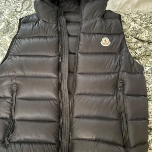 Svart dunväst från Moncler - Säljer en stilren svart dunväst från Moncler. Västen har en dragkedja framtill och två praktiska fickor med dragkedjor. Perfekt för kyligare dagar och ger en sportig look. Storlek 4