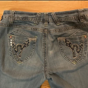 Blå jeans med broderade fickor - Snygga blå jeans med unika broderade detaljer på bakfickorna och dekorativa knappar. De har en bootcut-stil och är perfekta för en avslappnad look. 