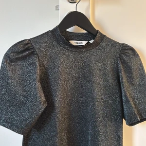 Glittrig topp från Kappahl - Säljer en glittrig topp från Kappahl i svart med korta puffärmar. Perfekt för en festlig look. Toppen har en rund halsringning och en elegant glansig finish. Är öppen på många prisförslag men max över 35 kr helst💞