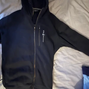 Svart tröja ifrån sail racing - Snygg svart jacka från Still Machine med dragkedja och huva. Perfekt för kyligare dagar med sin långärmade design och stilrena look.