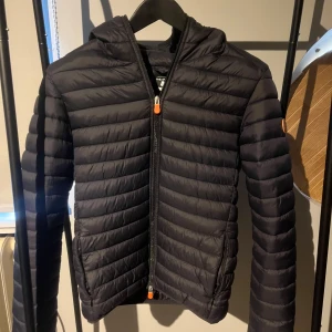 Jacka från Save The Duck. Storlek M - Mörk, lättviktsjacka med huva. Jackan har dragkedja fram och två fickor.