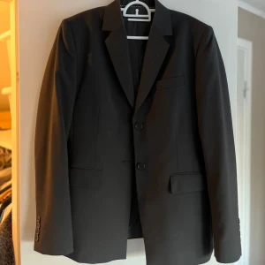 Svart kavaj från Turo Tailor - Elegant svart kavaj från Turo Tailor i storlek 38. Tillverkad av en blandning av polyester, ull och elastan för en bekväm passform. Kavajen har klassiska slag och knäppning med två knappar framtill. Perfekt för en stilren look. Ny pris 2000-3000