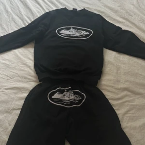 Corteiz set (shorts och tröja) Size XS/S - Säljer en svart tröja med ett coolt tryck av en byggnad på framsidan. Tröjan är tillverkad i en mjuk bomulls- och polyesterblandning och har långa ärmar. Perfekt för en avslappnad stil.