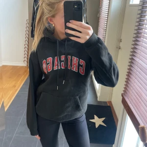 Svart hoodie med Chicago-tryck - Säljer en svart hoodie med texten 'CHICAGO' i rött och vitt på framsidan. Tröjan har en klassisk känguruficka och justerbar huva med dragsko. Perfekt för en avslappnad stil.