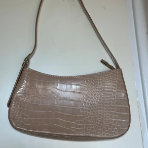 Beige axelväska med krokodil mönster - Snygg beige axelväska med krokodilpräglat mönster. Väskan har en dragkedja upptill och en justerbar axelrem. Perfekt för att ge en clean touch till din outfit 🫶🏾