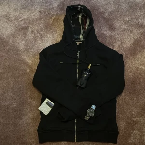 Svart hoodie från Burberry - Säljer en stilren svart hoodie från Burberry med dragkedja och klassiskt rutigt mönster i huvan. Perfekt för en avslappnad och trendig look. 🖤