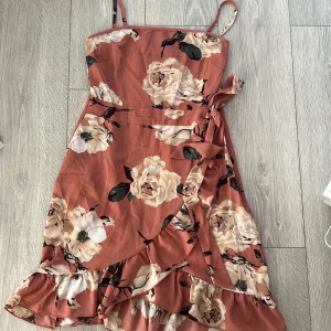 Blommig klänning med volangdetaljer - Säljer en vacker blommig klänning med stora vita blommor och gröna blad. Klänningen har smala axelband och en knytning i midjan. Volangdetaljer pryder nederdelen för en extra feminin touch.