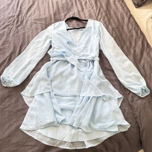 Ljusblå omlottklänning - Säljer en ljusblå omlottklänning med långa ärmar och volangdetaljer. Från nakd. Endast använd en kväll. Liten i storlek så passar S-M