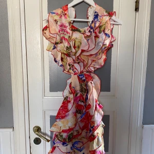 Färgglad blommig klänning med volanger - Säljer en färgglad kortklänning med blommigt mönster och volanger. Klänningen är ärmlös och har en öppen rygg, perfekt för sommarens fester. Den har en livfull mix av rött, blått och gult.