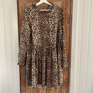 Leopardmönstrad klänning från Vero Moda Storlek M - Snygg leopardmönstrad klänning från Vero Moda med långa ärmar och en avslappnad passform. Perfekt för att sticka ut med stil. Klänningen har en rund hals och knappar vid ärmsluten för extra detalj. Använd 1 gång. 