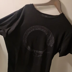 Svart t-shirt från Roberto Cavalli - Snygg svart t-shirt från Roberto Cavalli med ett glittrigt cirkelmönster på framsidan. Perfekt för att ge din outfit en lyxig touch. T-shirten har en normal passform och korta ärmar.