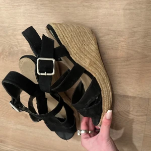 Kilklackar - Snygga svarta sandaler från din sko med kilklack och justerbara spännen, storlek 38. De har en öppen tå och är tillverkade i mocka med en flätad sula i botten. Använda endast 2 gånger.