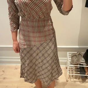 Rutig klänning med öppen rygg - Säljer en rutig klänning i beige, rött och svart med trekvartsärm och öppen rygg. Klänningen har en figurnära passform upptill. Min finaste klänning menr lite för liten för mig nu :(