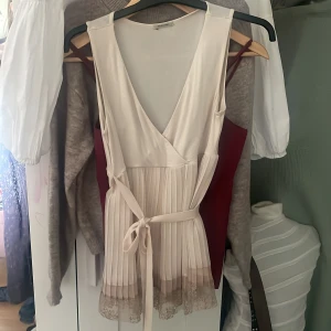 Vit omlott topp med spets från Intimissimi - Supersöt vit omlott topp från Intimissimi med plisserad kjol, knytband i midjan och spetsdetaljer längst ner. Toppen är ärmlös och har en v-ringad halsringning. Perfekt för dig som gillar romantiska och feminina detaljer.