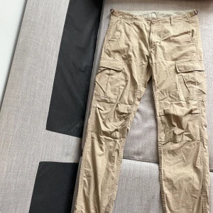 Beiga cargobyxor från Carhartt - Säljer ett par beiga cargobyxor från Carhartt med flera praktiska fickor på sidorna. Byxorna har normal passform och är tillverkade i slitstark bomull. Perfekta för dig som gillar funktionella och stilrena byxor.Helt oanvända 