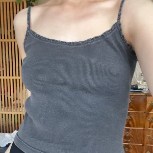 Mörkgrå ribbad linnetopp - Säljer en mörkgrå ribbad linnetopp med smala axelband och diskret spetskant upptill. Perfekt för varma dagar eller som lager under andra plagg. Enkel och stilren design.