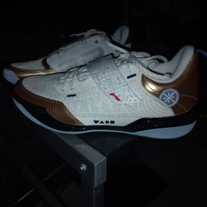 Way of wade 808 4 ultra "score champion" - Säljer ett par vita sneakers med guldiga detaljer och svarta inslag från Way of Wade. Skorna har snörning, rund tå och en platt sula med unikt mönster undertill. Ovandelen har textil och syntetmaterial med grafiska detaljer på kardborrebandet. Är även använd en gång, man ser lite damm som den har plockat upp. Kan torkas av enkelt.