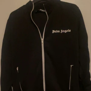 Svart track jacket från Palm Angels  - Säljer en äkta svart track jacket från Palm Angels med vita ränder längs ärmarna och vit dragkedja. Jackan har hög krage, två fickor med dragkedja och logga på bröstet. Perfekt för en sportig och trendig look.