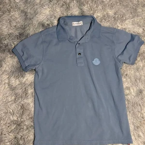 Ljusblå piké t-shirt från Moncler - Säljer en ljusblå piké t-shirt från Moncler med klassisk krage och två knappar framtill. T-shirten har korta ärmar och en broderad Moncler-logga på bröstet. Perfekt för en stilren och avslappnad look.