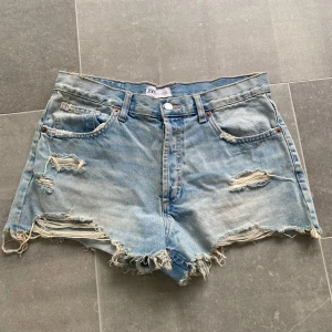 Ljusblå slitna jeansshorts från Zara - Ljusblå jeansshorts med fransiga kanter och slitningar på fram- och baksidan. Klassisk femficksmodell med knapp och dragkedja. Perfekta för sommaren och ger en avslappnad look.