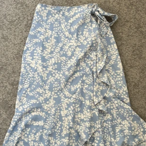 Blåvit omlottkjol från Gina Tricot - Säljer en blåvit omlottkjol från Gina Tricot med blommigt mönster och volangdetaljer längs kanten. Kjolen knyts i midjan och har en luftig känsla, perfekt för varmare dagar.