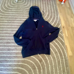 Mörkblå hoodie från U.S. Polo Assn. - Säljer en mörkblå hoodie från U.S. Polo Assn. med dragkedja och huva. Tröjan har två fickor framtill och en röd broderad logga på bröstet. Perfekt för en avslappnad stil och enkel att matcha med jeans eller joggers.