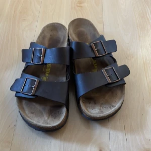 Mörkbruna sandaler från Birkenstock - Klassiska mörkbruna sandaler från Birkenstock med två breda remmar och justerbara metallspännen. Fotbädden är formad för extra komfort och sulan är platt. Perfekta för varma dagar och enkel att matcha med olika outfits.