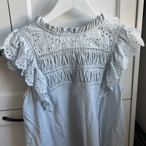 Ljusblå blus med spets från Holly & Whyte - Supersöt ljusblå blus från Holly & Whyte med broderade spetsdetaljer och volang vid axlarna. Blusen är ärmlös och har en romantisk känsla med fina hålbroderier och rynkade partier upptill.