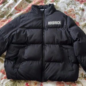 Snygg svart pufferjacka från Hoodrich.  - Säljer en snygg svart pufferjacka från Hoodrich i storlek M, kan användas för även någon med storlek S. Jackan har dragkedja framtill, hög krage och stora logotyper både fram, en liten på armen och bak. Två fickor på sidorna och en rund patch på ärmen. Perfekt för kyliga dagar. Säljs pga inte använder. 