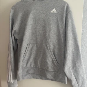 Grå hoodie från Adidas med vita ränder - Säljer en grå hoodie från Adidas med klassiska vita ränder längs ärmarna och över ryggen. Tröjan har magficka och Adidas-logga på bröstet. Perfekt för en sportig och avslappnad stil.