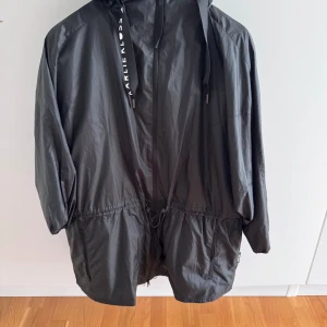 Svart vindjacka från Adidas  - Säljer en svart vindjacka från Adidas  med huva och dragsko i midjan. Jackan har långa ärmar, dragkedja framtill och snörning i huvan. Perfekt för blåsiga dagar.