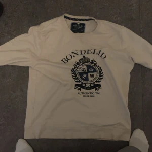 Vit långärmad tröja från Bondelid - Säljer en vit långärmad tröja från Bondelid med tryckt emblem och text på bröstet. Tröjan har rund halsringning och klassisk passform. Perfekt för dig som gillar stilrena och tidlösa plagg.