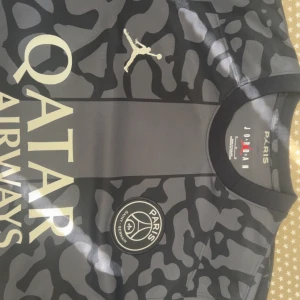 PSG x Jordan svart fotbollströja kvitto finns - Snygg svart PSG fotbollströja i samarbete med Jordan. Tröjan har ett unikt mönster, guldiga detaljer och Paris Saint-Germain-logga på bröstet. Qatar Airways som sponsortryck och klassisk rund hals. Perfekt för fotbollsfans! 