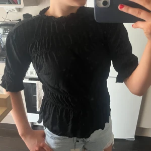 Svart blus med rynkade detaljer - Säljer en svart blus med rynkade partier och lätt volang vid halsringningen. Blusen har trekvartsärm och en figurnära passform i midjan som ger en snygg siluett. Perfekt att styla med jeans eller kjol.